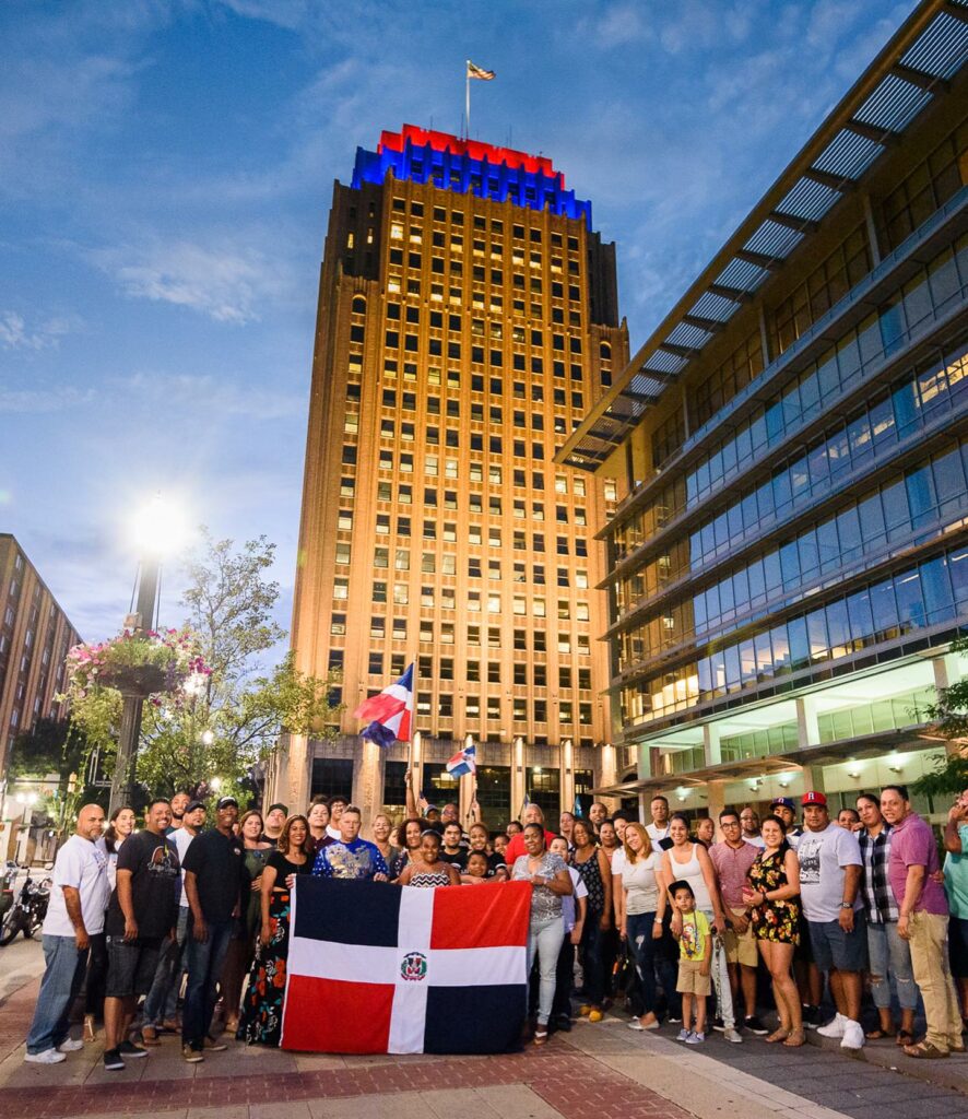 A crowd of people holding Dominican flags pose facing the camera. The PPL tower in Allentown is in the background against the early evening sky.